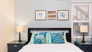 Ein Schlafzimmer mit einem Bett, zwei Nachttischen, einem Fenster mit Jalousien und drei gerahmten Bildern an der Wand.