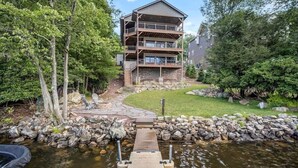 A multi-story building with a stone facade, a wooden deck leading to a dock over water, and a well-maintained lawn. - Water's Edge!  11 Bdrm, 5.5 Bath, Lakefront, Dock, Sleeps 35, Hot Tub, IR Sauna (Lake Harmony)