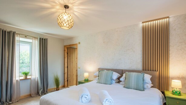 A neatly made bed with two rolled towels, a wooden door, a window with curtains, and a wall-mounted light fixture. - Cooriedoon by Interhome (Hopeman)