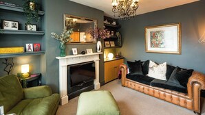 A living room with a fireplace, a green armchair, a brown leather sofa, and a chandelier. - Historic 2BR Home - Garden, BBQ, Parking (Greater London)