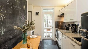 A modern kitchen with white cabinets, a blackboard with chalk drawings, a dining table set with plates and cutlery, and a view of the outdoors through a glass door. - Historic 2BR Home - Garden, BBQ, Parking (Greater London)
