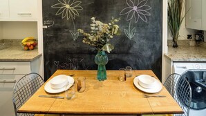 A dining table set with plates, cutlery, and a vase with flowers. A chalkboard with floral drawings and the text "Enjoy your stay!" is visible in the background. - Historic 2BR Home - Garden, BBQ, Parking (Greater London)