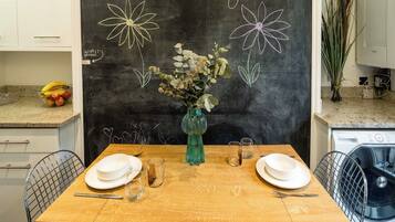 A dining table set with plates, cutlery, and a vase with flowers. A chalkboard with floral drawings and the text "Enjoy your stay!" is visible in the background.