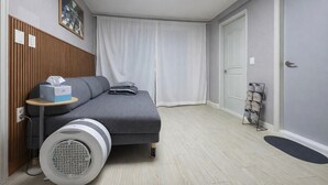 Una habitación de hotel moderna y compacta, con un sofá gris, una mesita con una caja de pañuelos, un ventilador de pie y un zapatero cerca de la puerta.