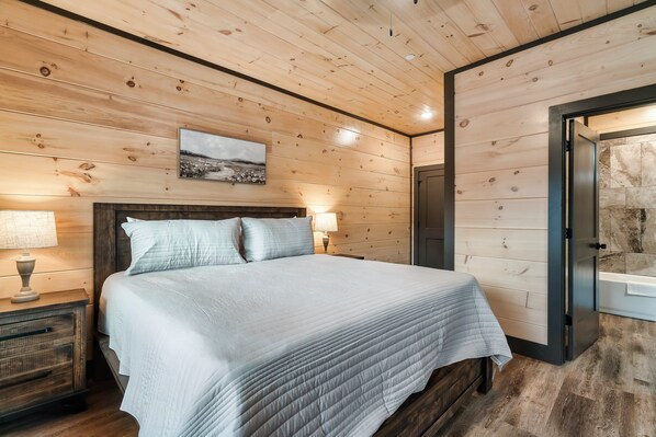 A bedroom with a wooden ceiling, a bed with a white quilt, a nightstand with a lamp, and a painting on the wall. - NEW! 12BR, Pool, Hot Tub, Sleeps 26 (Sevierville)