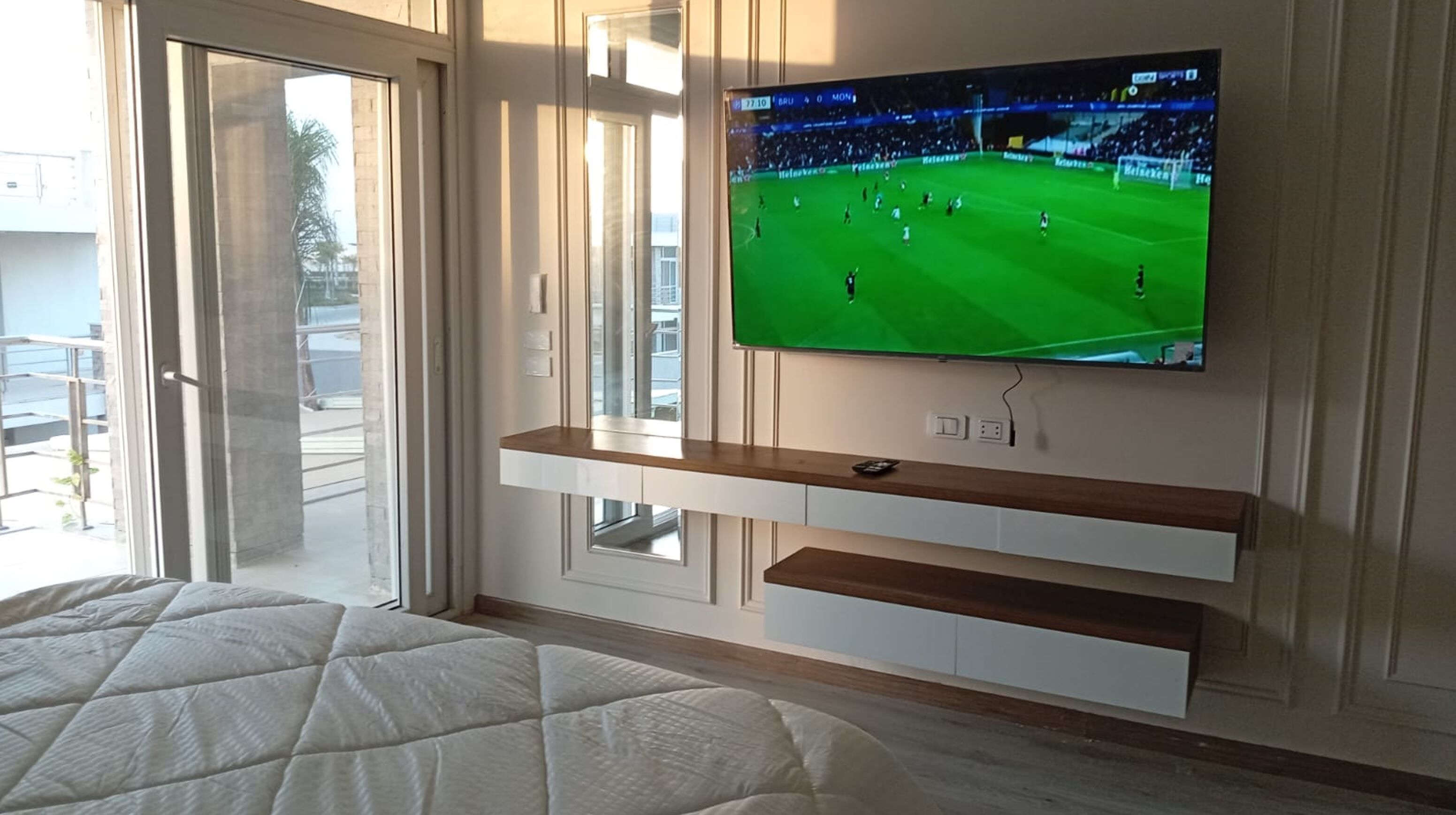 Ein modernes Wohnzimmer mit einem großen Flachbildfernseher an der Wand, einer Glasschiebetür, die auf einen Balkon führt, und einem Bett mit einer gesteppten Tagesdecke.