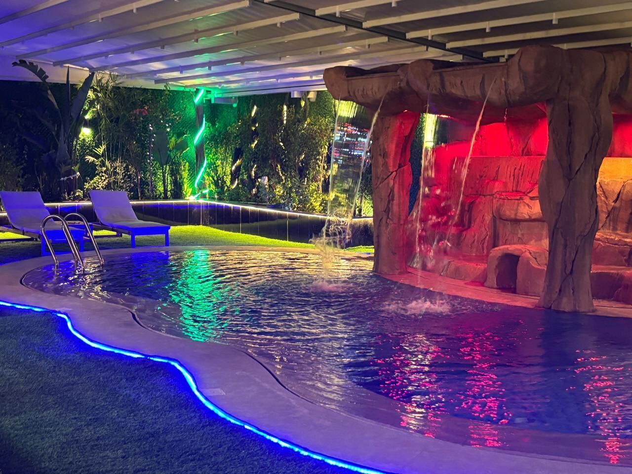 A swimming pool with a waterfall feature, surrounded by lounge chairs and a rock formation.