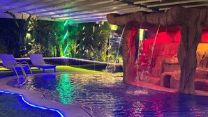 Una piscina con una cascada, rodeada de sillas de descanso y una formación rocosa.