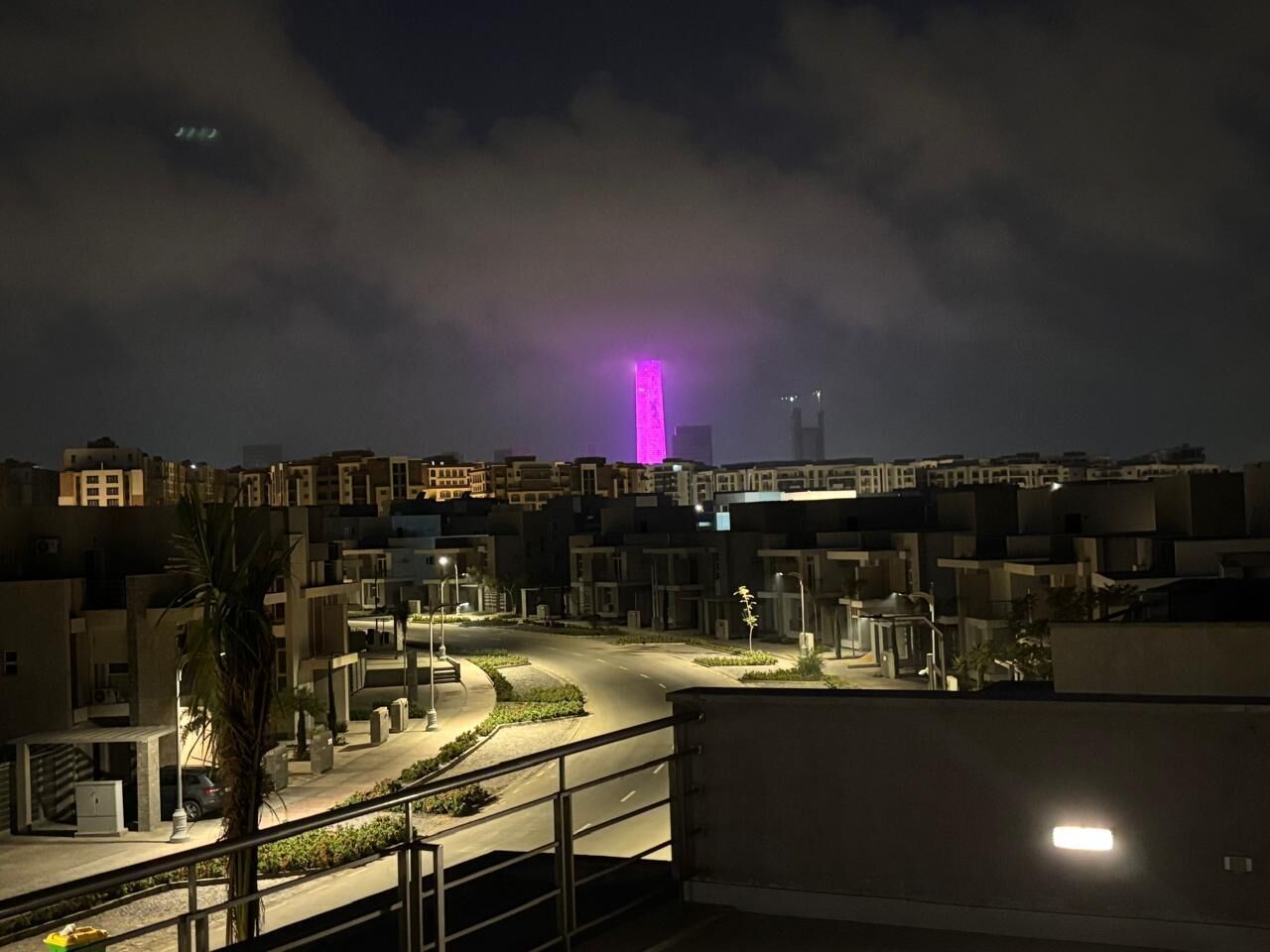 A nighttime view of a residential area with buildings, a curved road, and a pink-lit tower in the distance.