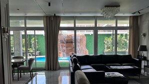 A spacious living room with a large black sectional sofa, a coffee table, and a dining area with a table and chairs. Large windows provide a view of a swimming pool and a rock formation outside.