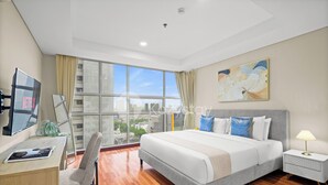 A modern hotel room with a large bed, a desk with a laptop, a chair, a bedside table with a lamp, and a large window offering a city view. - Suvin by Kozystay - Pondok Indah (Jakarta)