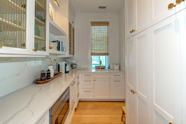 A modern kitchen with white cabinetry, a marble countertop, and a window with a woven blind. - Brand New Custom Home~ Oceanfront, Pool, Hot Tub, Elevator (Isle Of Palms)