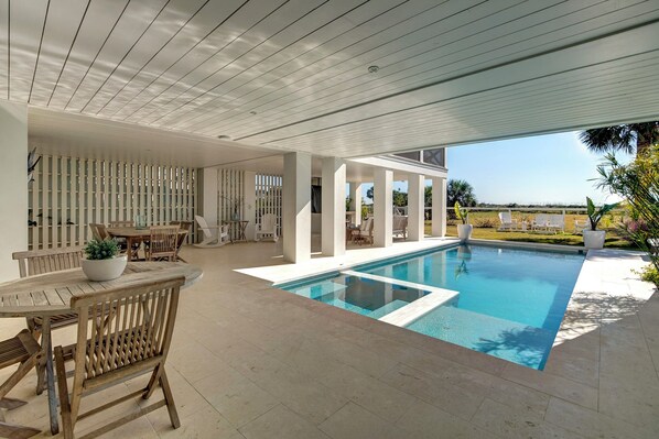 A modern outdoor pool area with a wooden deck, seating, and a view of the surrounding landscape. - Brand New Custom Home~ Oceanfront, Pool, Hot Tub, Elevator (Isle Of Palms)