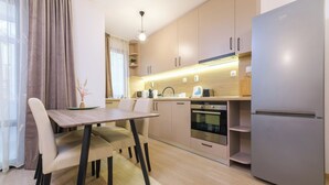 A modern kitchen with a dining area, featuring a built-in oven, a refrigerator, and a wooden dining table with chairs. - Comfort 1 Bedroom App (Plovdiv)