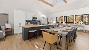 A modern kitchen with white cabinets, a central island with stools, a dining area with a large table and chairs, and large windows with autumn foliage views.