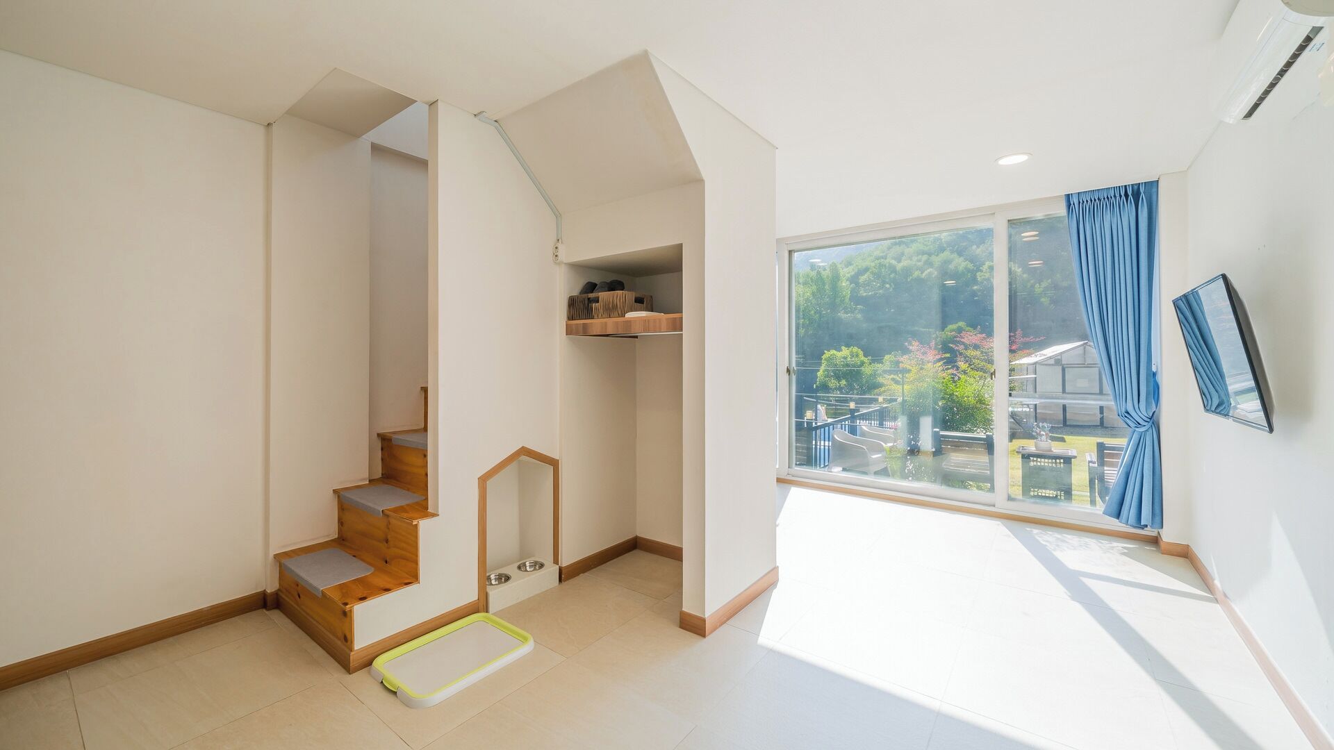 A spacious room with a wooden staircase, a pet bed, and a large window with a view of greenery.