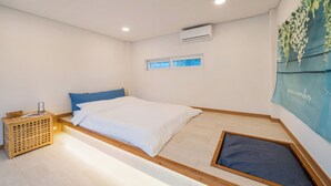 A bedroom with a bed, a wooden bedside table with a lamp, an air conditioning unit, a window with a view, and a blue wall hanging.