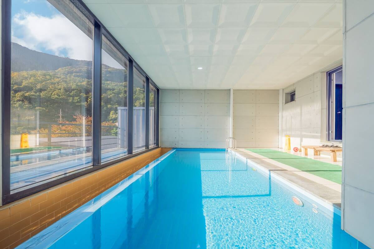 A swimming pool with clear blue water, large windows, and a view of a forested area.