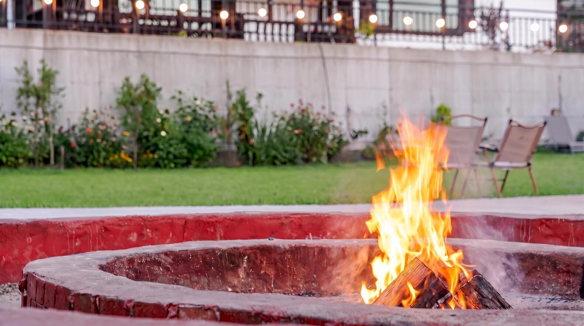 A circular fire pit with flames, surrounded by a red border, in an outdoor setting with chairs and a concrete wall in the background.