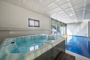 Una piscina interior moderna con jacuzzi, grandes ventanales y vistas a zonas verdes.
