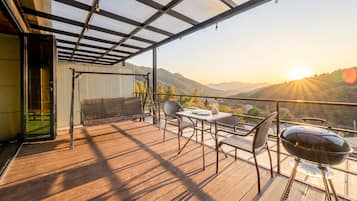 Un balcón con piso de madera, una mesa, sillas, una parrilla y vistas a las montañas.