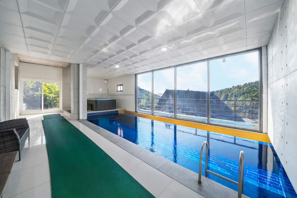 An indoor swimming pool with clear glass walls, a green mat, and a view of a forested hill.
