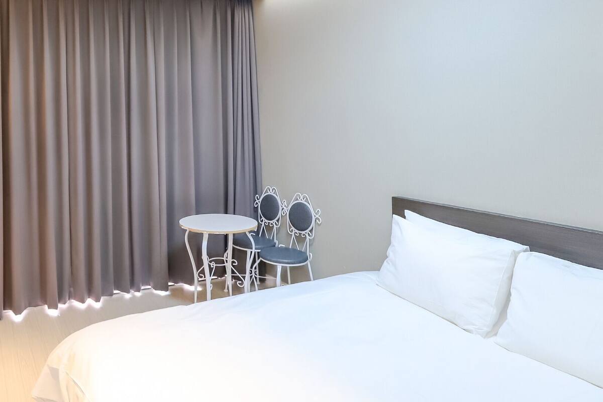 Una habitación de hotel con una cama, una mesita redonda pequeña y dos sillas, y una cortina.