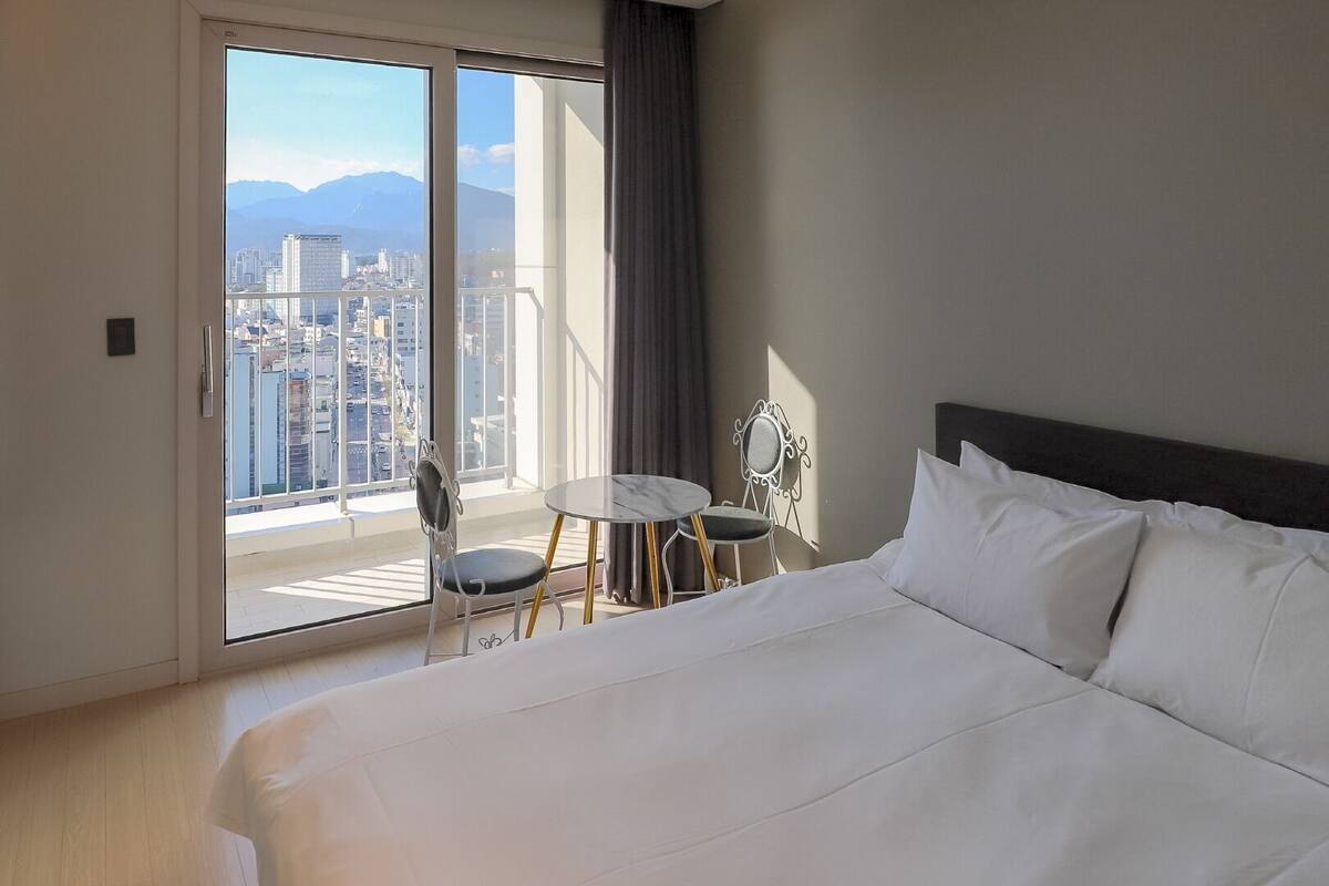 Habitación de hotel con una cama grande, una mesita pequeña, dos sillas y vistas a un paisaje urbano a través de un ventanal amplio.