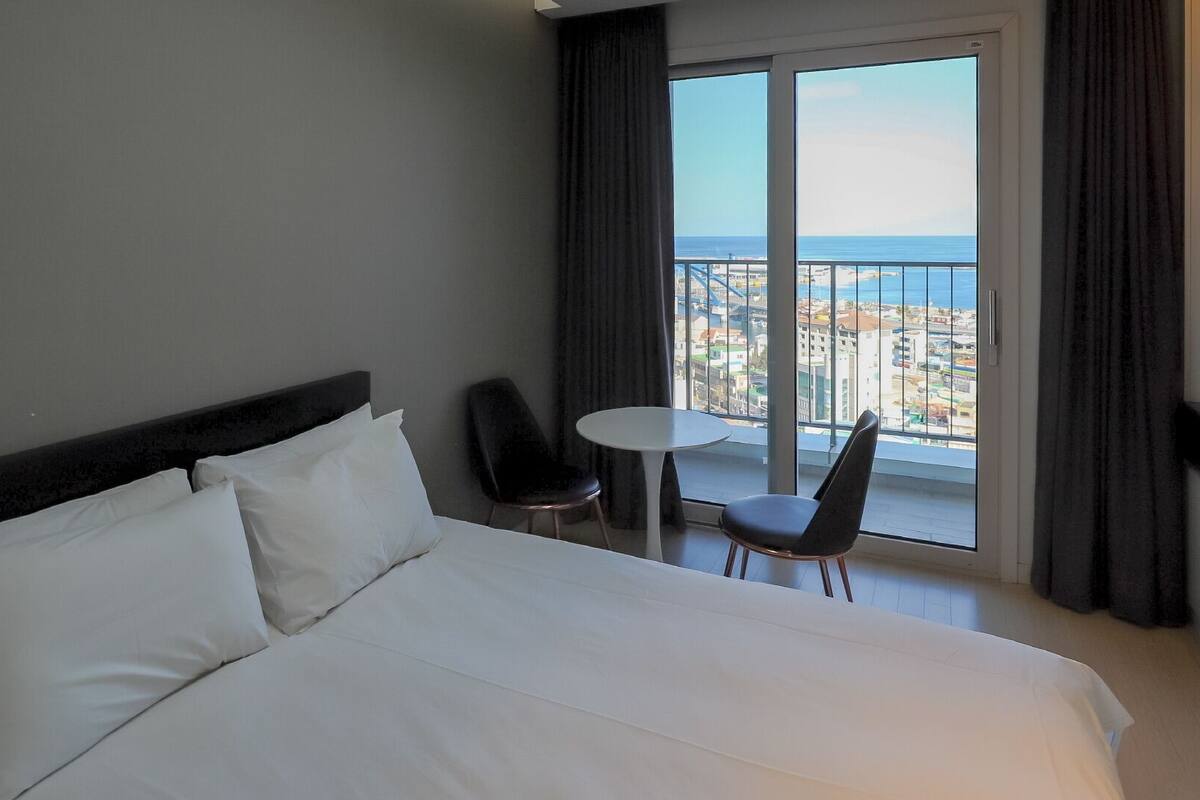 Habitación de hotel con una cama grande, dos sillas, una mesita y vistas a la ciudad y al mar a través de amplios ventanales.
