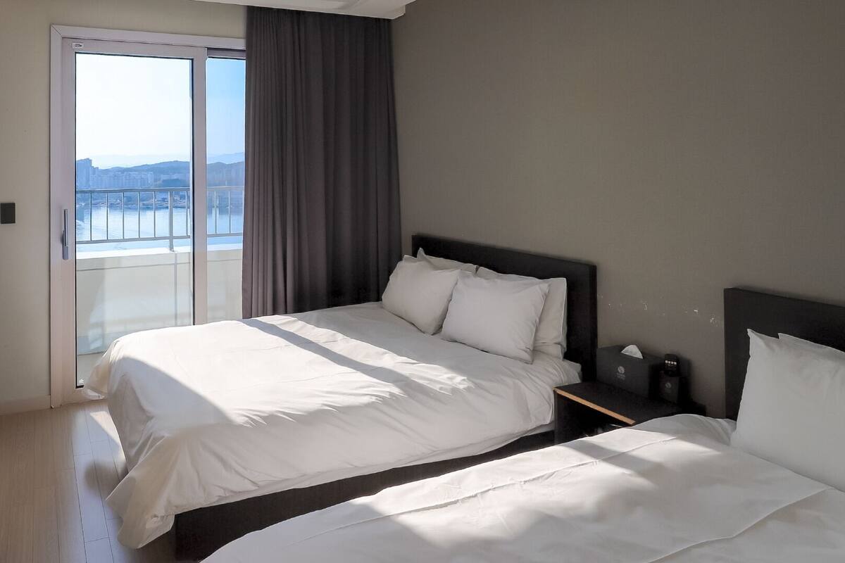 Habitación de hotel con cama, mesita de noche y vista a un paisaje urbano a través de un amplio ventanal.