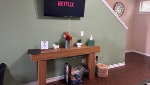A living room with a TV mounted on the wall displaying 'NETFLIX', a wooden console table with a vase, a clock, and a tissue box.