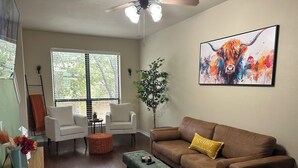 A living room with a brown sofa, two white armchairs, a green ottoman, and a coffee table. There is a painting on the wall, a plant, and a window with blinds.