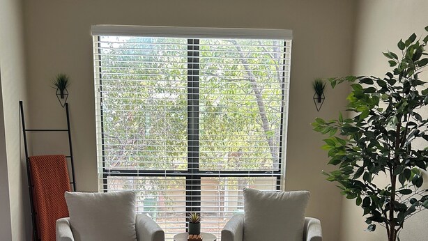 A living room with two white armchairs, a round ottoman, a small table with a plant, and a large window with blinds.