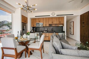 A modern kitchen with wooden cabinets, a stainless steel refrigerator, and a built-in microwave. There is a dining area with a glass table and chairs, and a gray sofa. - D'Vine Paradise - Luxe 2BR/2BA Retreat/Rooftop Pool & Spa (Puerto Vallarta)