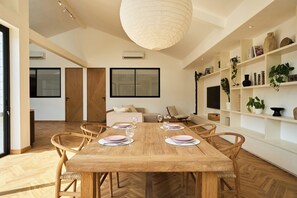 A dining area with a wooden table set for four, surrounded by wicker chairs. There is a large window above the table, and a white pendant light hangs from the ceiling. - White Villa by Betterplace (Bengkel)
