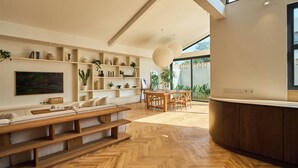 A modern living room with a herringbone wooden floor, a wooden dining table, and built-in shelving with plants and books. - White Villa by Betterplace (Bengkel)