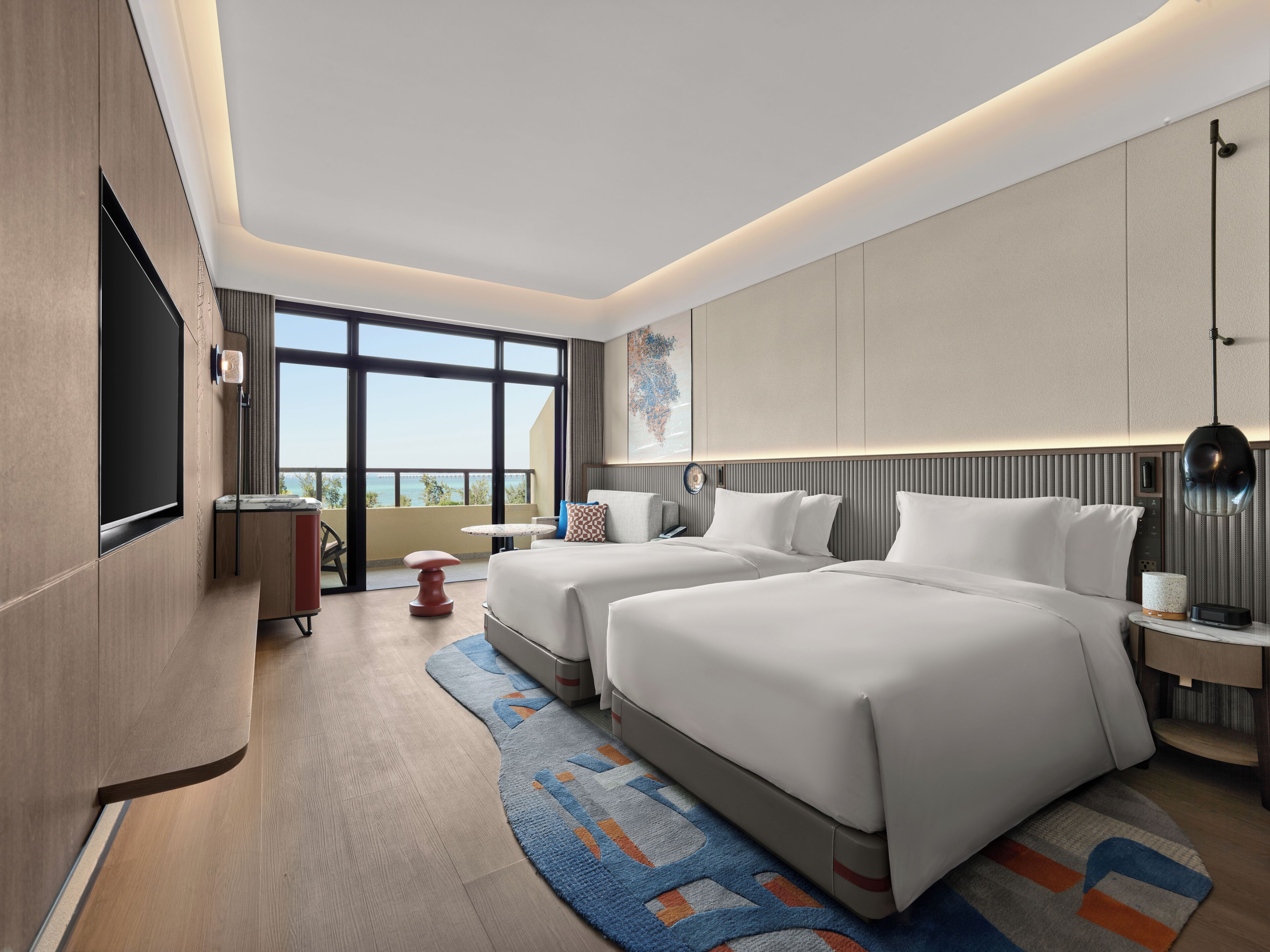 A modern hotel room with two beds, a large window, a flat-screen TV, and a view of the ocean.