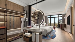 Una habitación de hotel moderna con una cama grande, un espejo redondo, un tocador con lavabo, un espacio de descanso y un amplio ventanal con vistas.