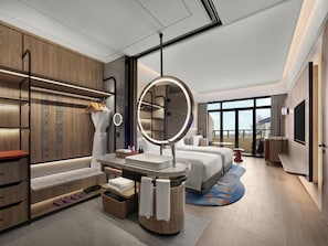Una habitación de hotel moderna con una cama grande, un espejo redondo, un tocador con lavabo, un espacio de descanso y un amplio ventanal con vistas.