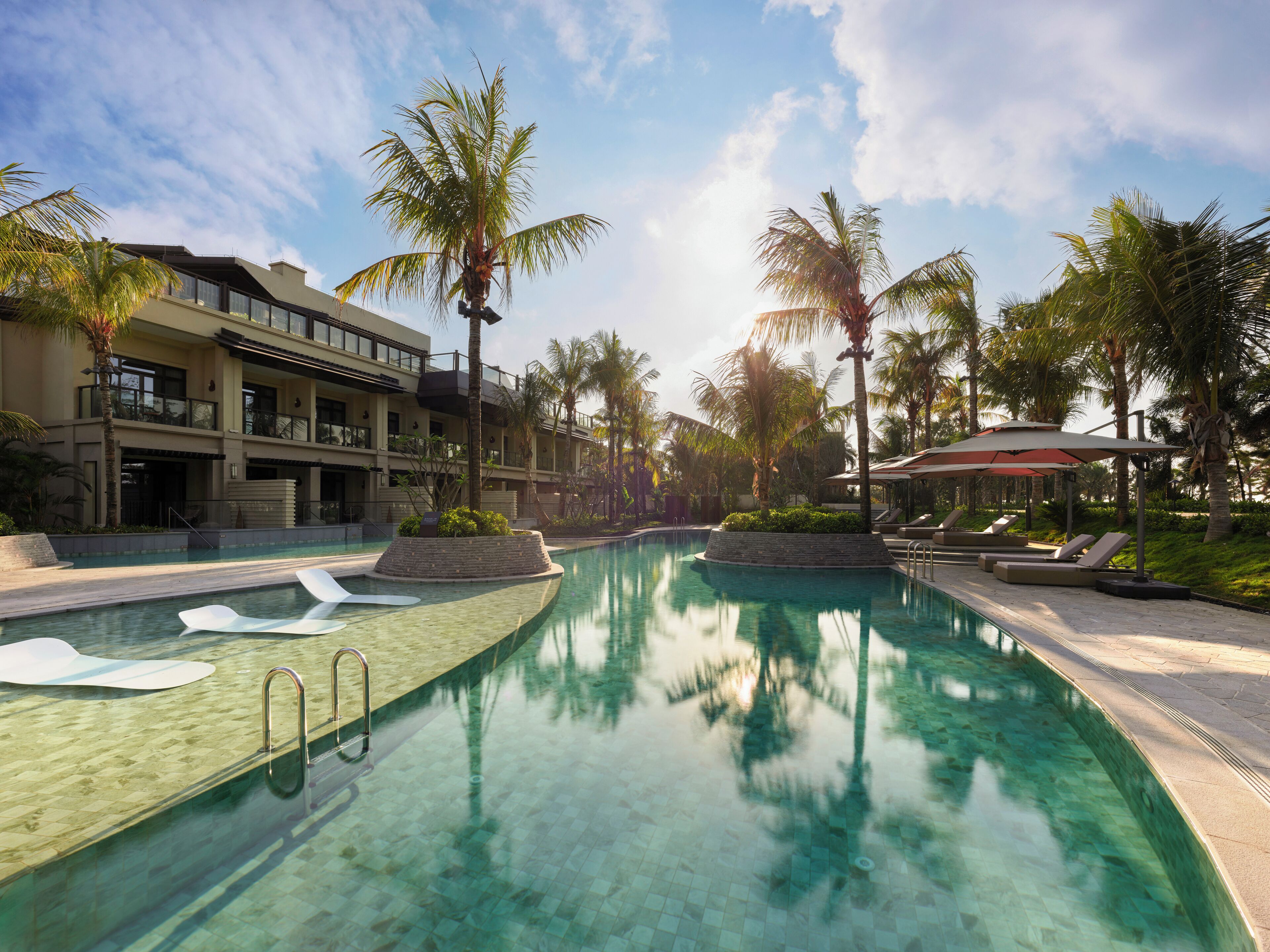 A clear swimming pool with lounge chairs and palm trees, surrounded by a resort building.