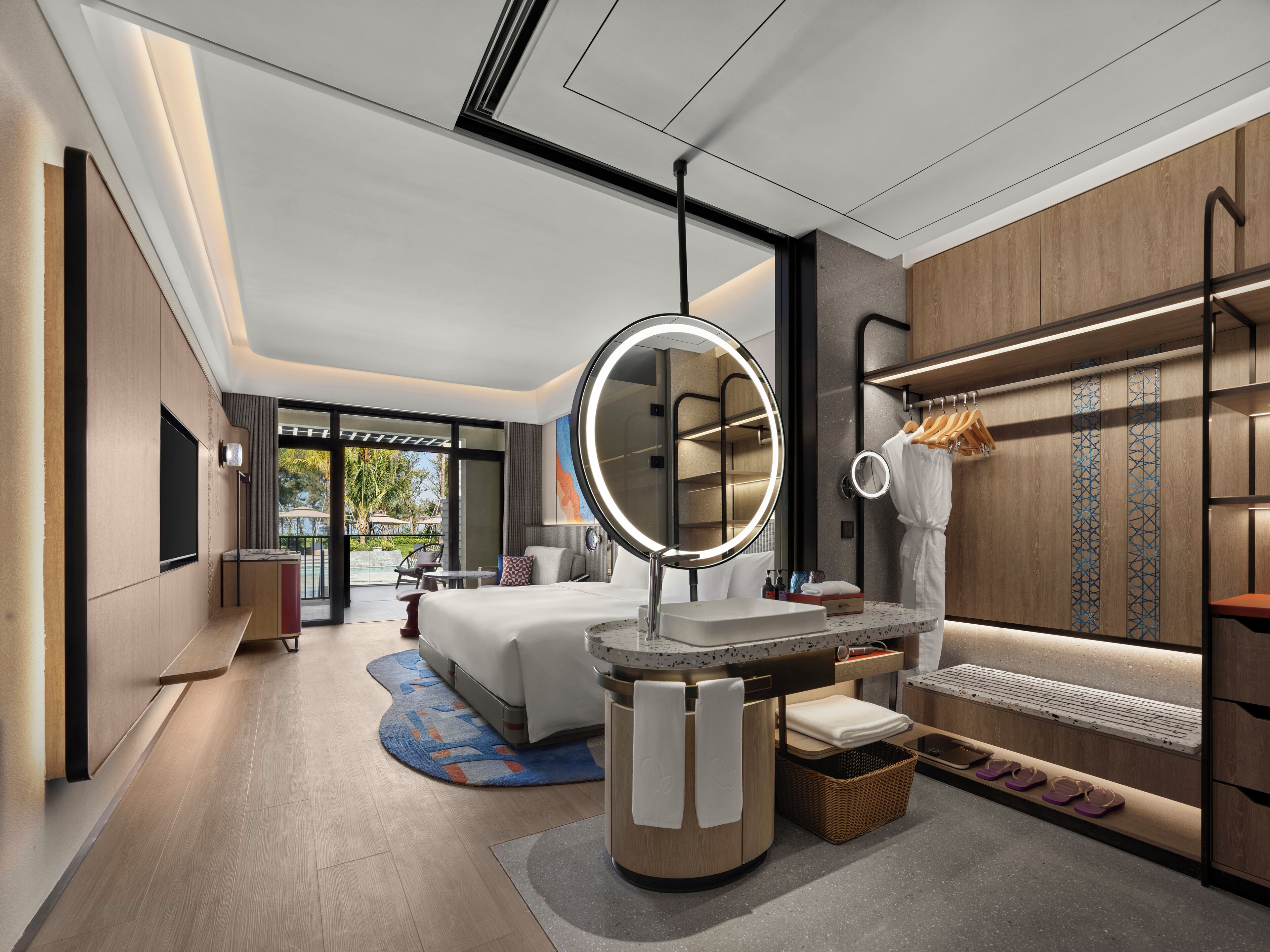 Une chambre d’hôtel moderne avec un grand lit, un miroir rond, une coiffeuse avec un lavabo et une vue sur l’extérieur.