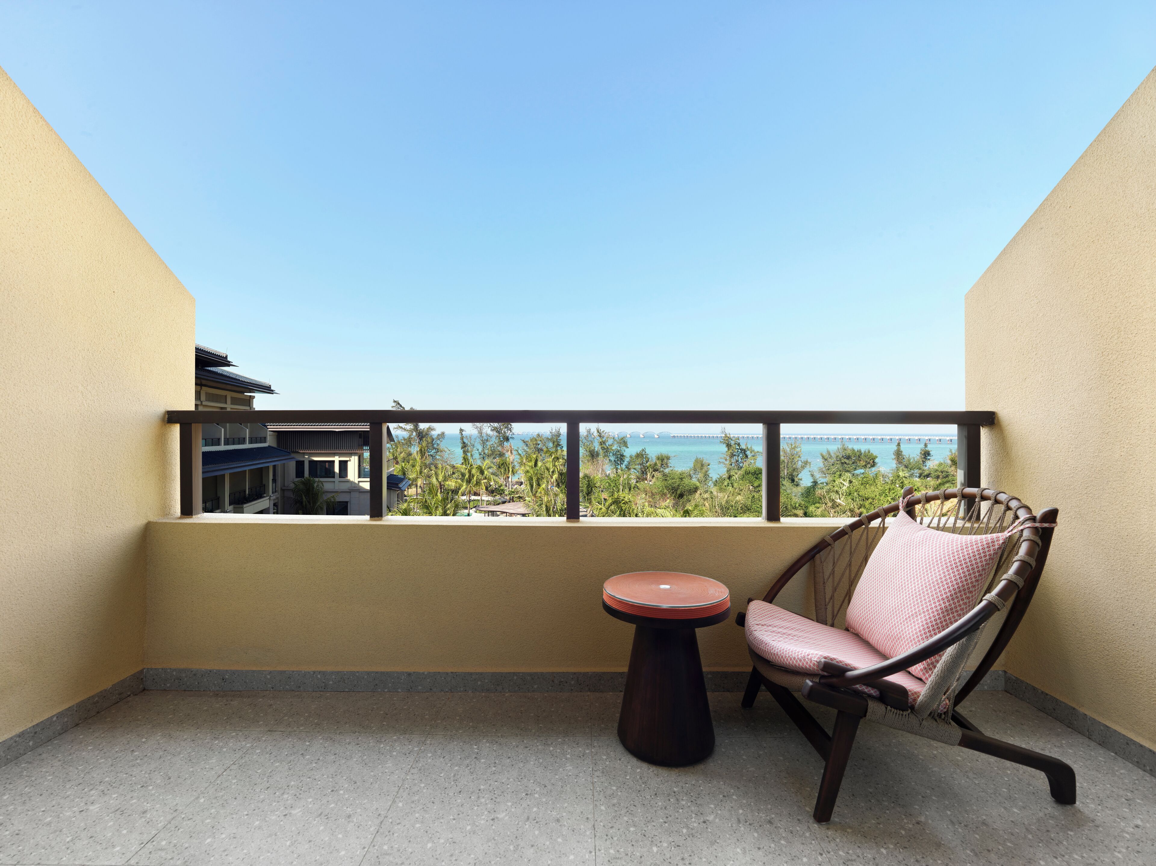 A balcony with a chair and a small table, overlooking a view of a bridge and greenery.