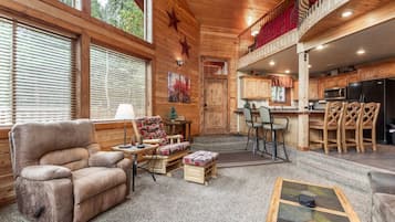 Un acogedor interior de cabaña con un sillón mullido, una pequeña mesa con lámpara, un sofá, un comedor con taburetes altos y una cocina con armarios de madera y un refrigerador negro.