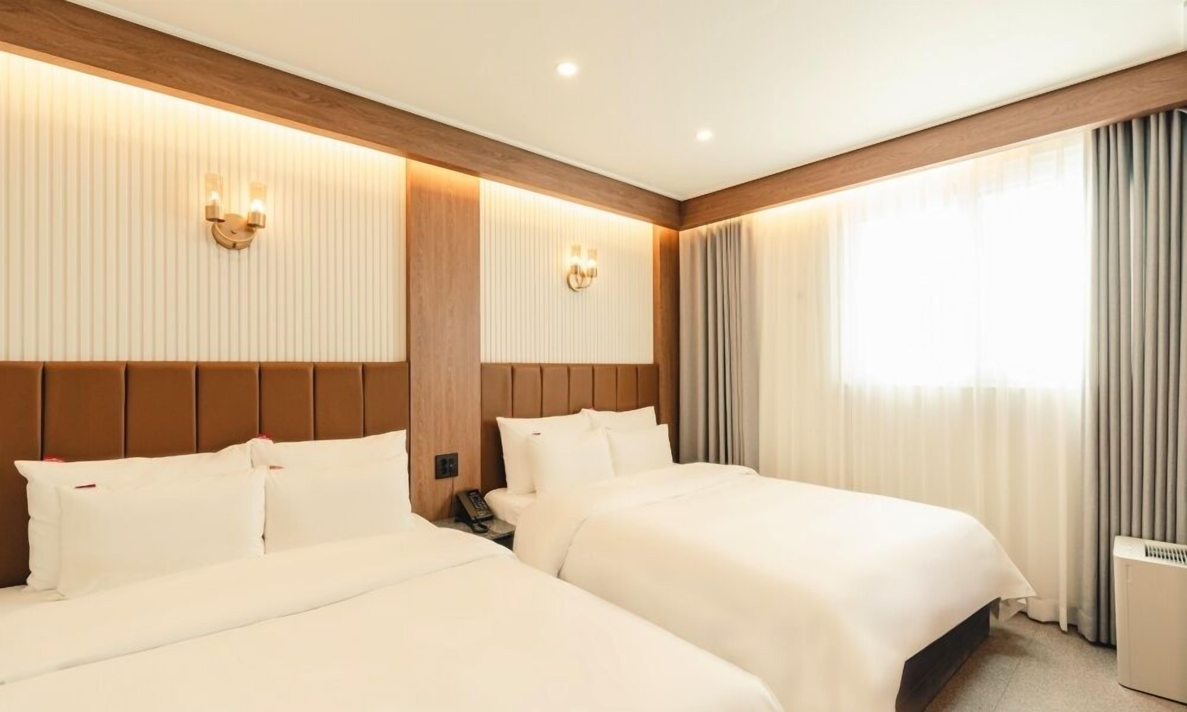 A hotel room with two beds, a wooden headboard, wall-mounted sconces, and a large window with curtains.