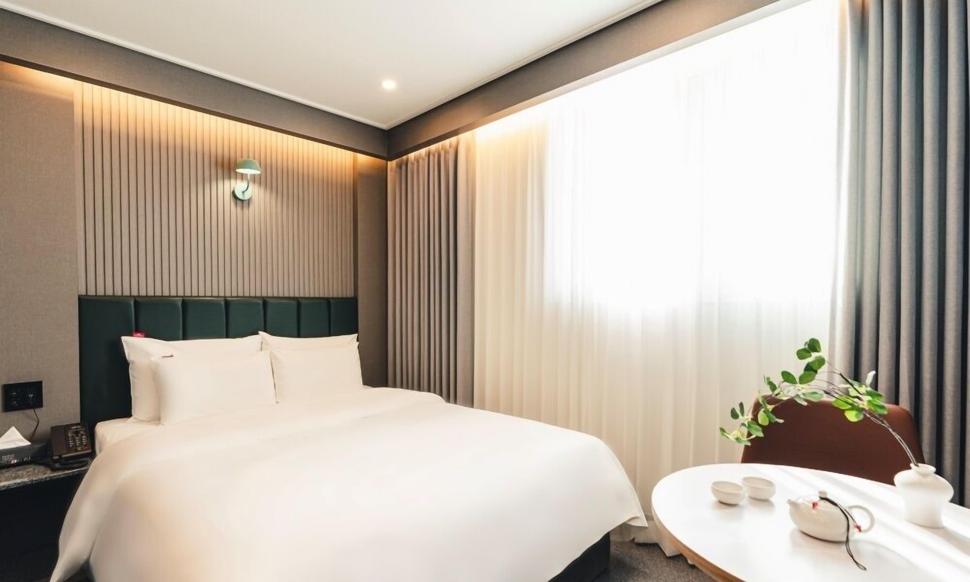 A hotel room with a large bed, a bedside table with a lamp, a small table with cups and a teapot, and a window with curtains.
