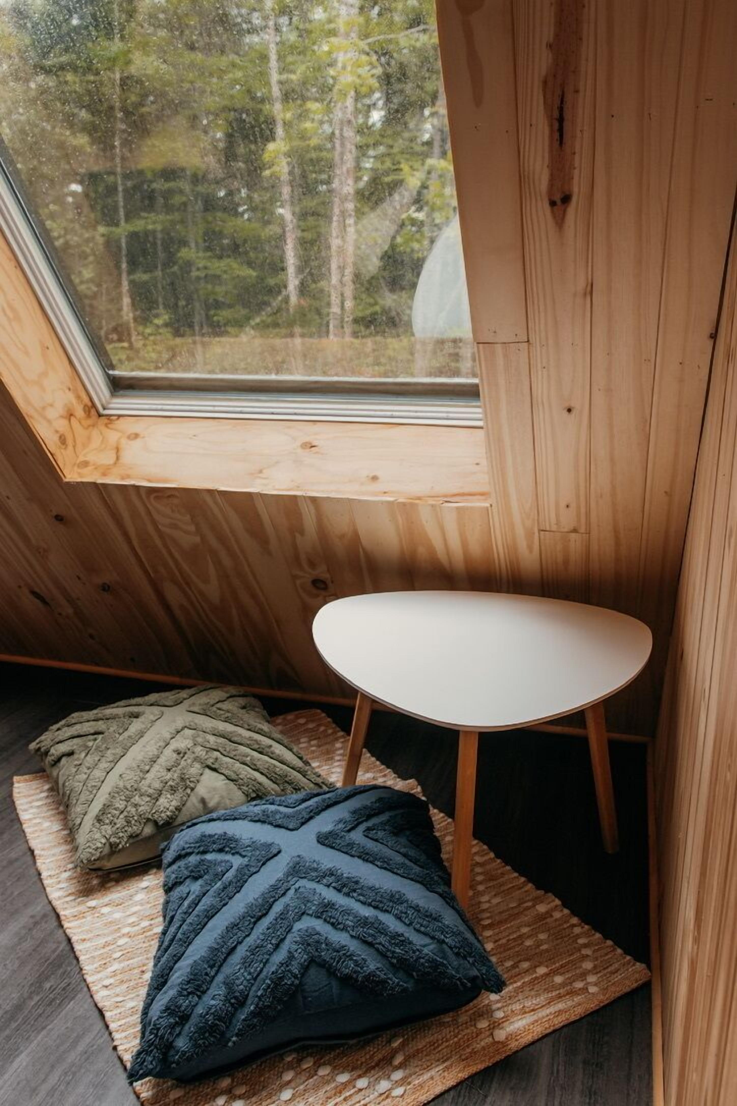 Een knus, klein kamertje met een schuine plafond, een witte tafel en drie gestructureerde kussens op een geweven vloerkleed.