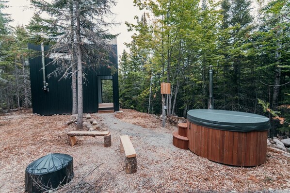 Una cabaña rústica con jacuzzi, bancos de madera y un área cubierta, rodeada de árboles.