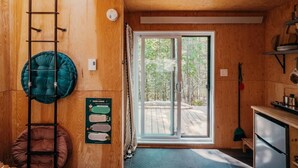 A cozy cabin interior with a wooden ceiling, a kitchenette, a seating area with cushions, and a large sliding glass door leading to an outdoor deck. - Domaine à Ciel Ouvert (La Malbaie)