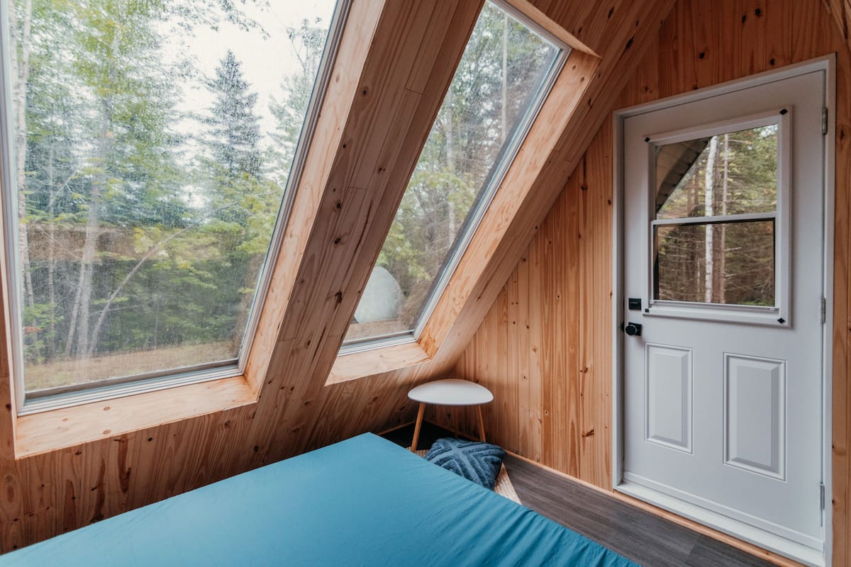 A room with a sloped ceiling, a bed, a door, and large windows offering a view of trees.