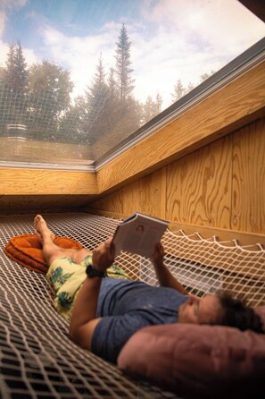 A person lying on a hammock in a wooden room with a view of trees through a window. - Domaine à Ciel Ouvert (La Malbaie)